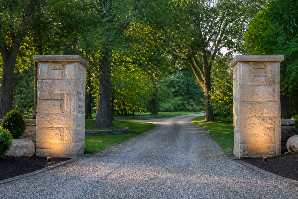 Grand entrance to an estate property featuring large natural stone pillars, a gravel driveway, and lush green trees in summer.