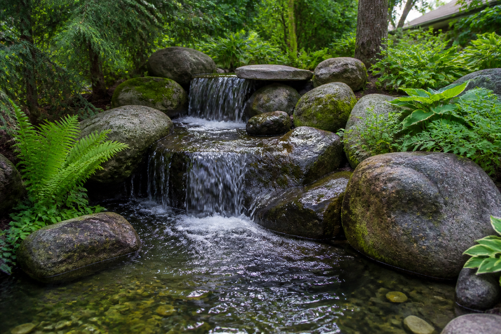 A close-up of a backyard waterfall with water flowing over large, weathered granite boulders into a pond surrounded by lush green garden plants.
